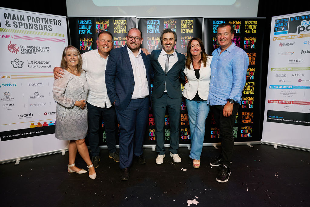 Leicester Comedy Festival Business Partners Club members Red Monkey Play & ER Recruitment stand on stage with Big Difference Company CEO & comedian Patrick Monahan in front of a set of business partners banners